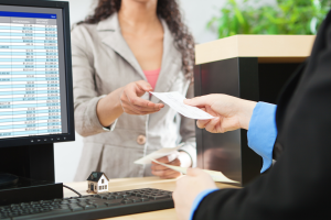 Picture of a person receiving a check from a bank teller. On the desk is a small model of a home. This represents the concept of accessing assets in a north carolina trust.