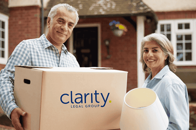 Photo of a man carrying a box and a woman carrying a lamp as they move into a new home. Represents updating wills and trusts after moving for NC residents.