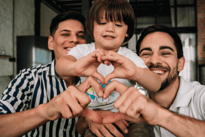 Image of a gay couple with their child. They're forming hearts with their hands. This illustrates who benefits from Estate planning for LGBTQ+ families