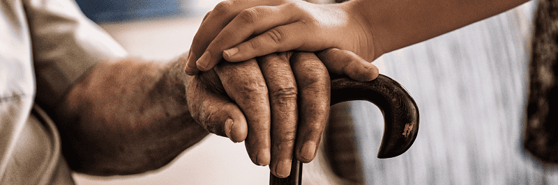 Photo of an elderly person with their hand on a crutch and a loved one holding their hand. The photo humanizes the idea of needing medicaid.