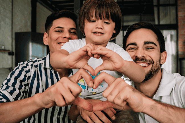 Image of a gay couple with their child. They're forming hearts with their hands. This illustrates who benefits from Estate planning for LGBTQ+ families